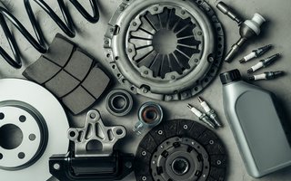 Verschiedene Autoersatzteile und Zubehörteile, darunter Bremsscheibe, Bremsbeläge, Kupplungsteile, Zündkerzen, Lager, Federn und ein Motorölbehälter, auf grauem Untergrund angeordnet.