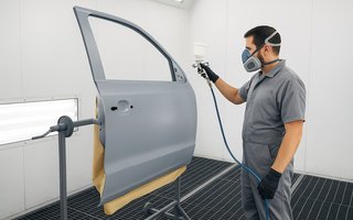 Lackierer mit Atemschutzmaske sprüht Farbe auf eine Autotür in einer Lackierkabine